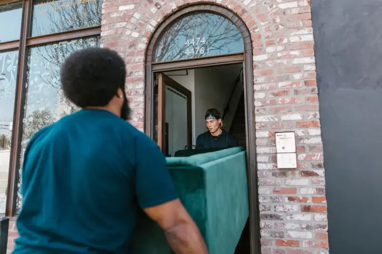 Dos operarios de mudanzas pasan un sofá verde por la puerta de un edificio de ladrillo